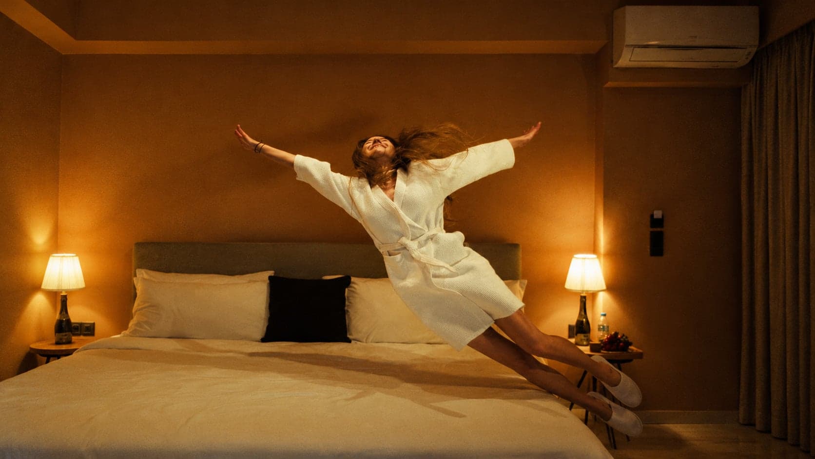A woman in a white robe leaps onto a hotel bed in warm lamplight
