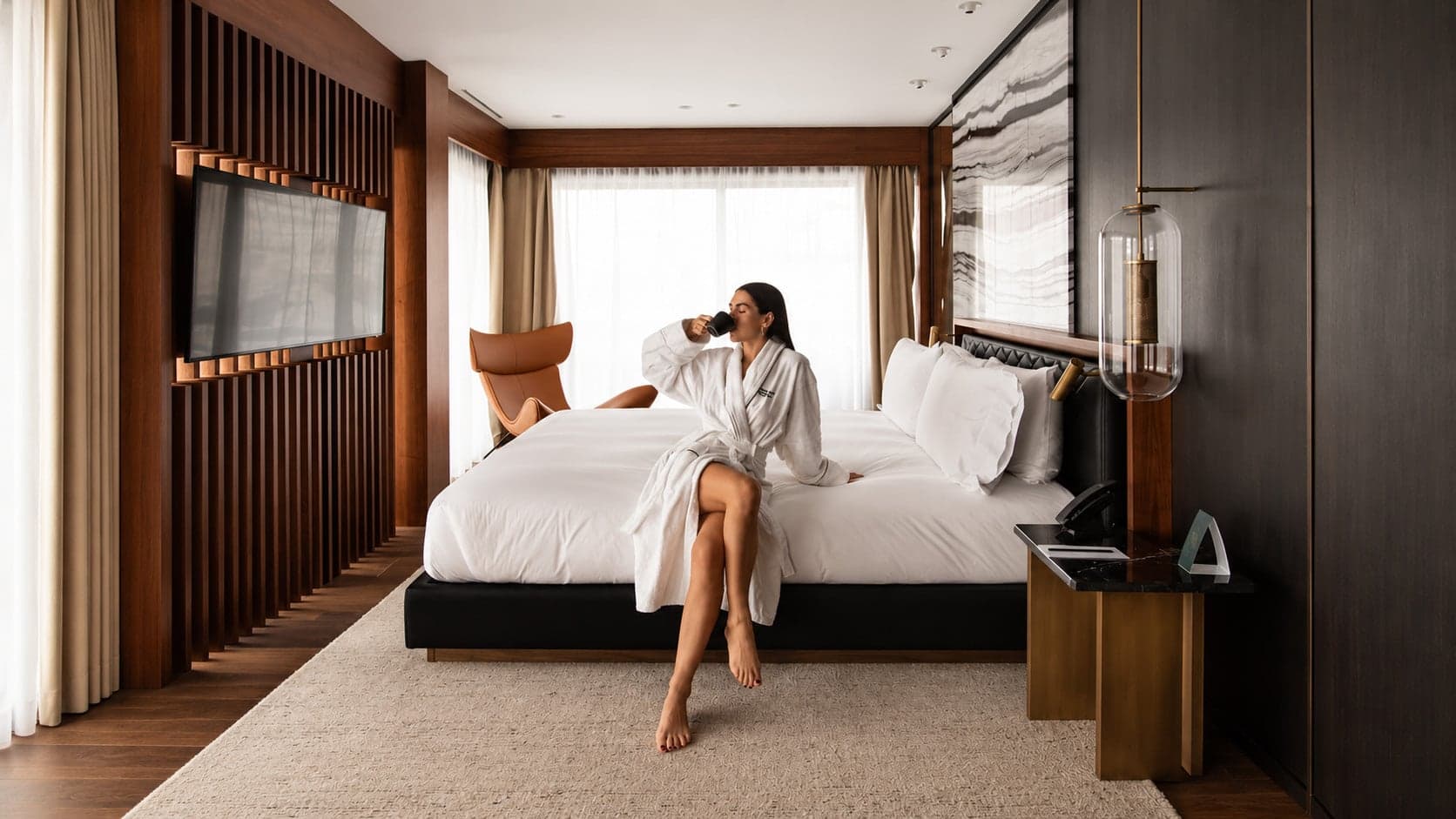A woman in a robe sipping from a mug on the edge of a hotel bed